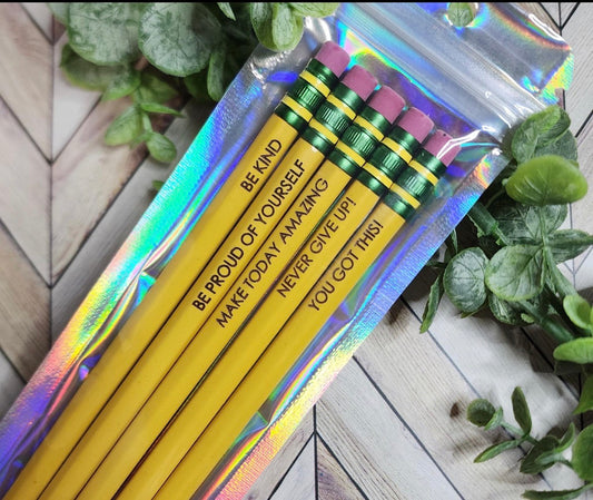 A set of five yellow pencils in a clear holographic zip pouch, placed on a light wood surface with green leaves around it. The pencils have pink erasers, green ferrules, and black text that reads: 'BE KIND', 'BE PROUD OF YOURSELF', 'MAKE TODAY AMAZING', 'NEVER GIVE UP!', and 'YOU GOT THIS!'.